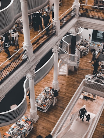 A multi-level bookstore features wooden floors, elegant white pillars, and winding staircases. People are browsing through books placed on neatly arranged tables and shelves. The architecture combines classic and modern elements, with open spaces and artistic railings.