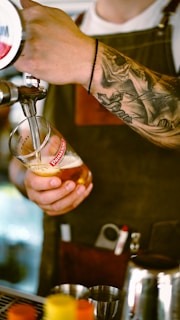A bartender with a tattoo on their arm is pouring beer from a tap into a glass. The glass has the brand name Prima filed on it. The bartender is wearing a dark apron over a white shirt.