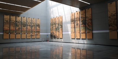 A large, modern hall featuring a series of tall, vertical paintings depicting traditional landscape scenes. The paintings are evenly spaced along two walls and are illuminated by natural light streaming through a skylight. The floor is reflective and made of dark tiles, adding a sense of spaciousness.