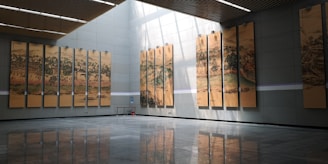 A large, modern hall featuring a series of tall, vertical paintings depicting traditional landscape scenes. The paintings are evenly spaced along two walls and are illuminated by natural light streaming through a skylight. The floor is reflective and made of dark tiles, adding a sense of spaciousness.