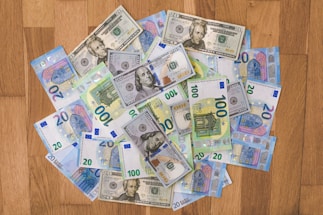 A collection of various denominations of U.S. dollar and Euro banknotes is scattered on a wooden surface. The banknotes include 20 and 100 dollar bills as well as 20 and 100 euro bills.