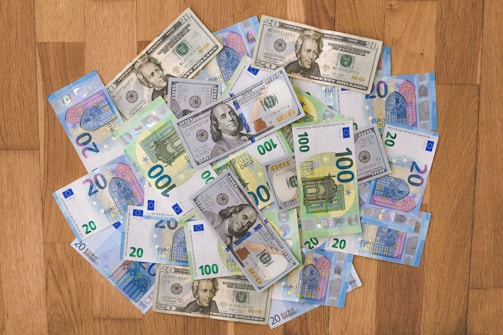 A collection of various denominations of U.S. dollar and Euro banknotes is scattered on a wooden surface. The banknotes include 20 and 100 dollar bills as well as 20 and 100 euro bills.