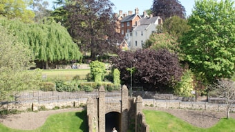 A lush garden with various trees including willow and other deciduous types surrounds a historic stone archway. Beyond the trees, several charming residential houses with brick and stucco exteriors stand in the background. The garden is enclosed by a decorative metal fence, and there is a well-manicured lawn.