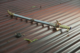 A decorative sword with intricate gold detailing is lying on a corrugated metal surface. The sheath is mostly light blue, adding contrast to the gold embellishments. Small pieces of debris and green leaves are scattered around the sword.