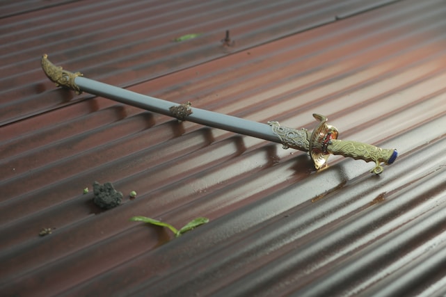 A decorative sword with intricate gold detailing is lying on a corrugated metal surface. The sheath is mostly light blue, adding contrast to the gold embellishments. Small pieces of debris and green leaves are scattered around the sword.