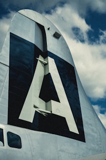 The image features the vertical stabilizer of an aircraft, prominently displaying a large letter 'A' against a dark background. The structure appears metallic with visible rivets, set against a partly cloudy sky.