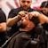 A barber is carefully trimming the hair of a customer who is leaning back in a barber's chair. The focus is on the precision and attention to detail in grooming the customer's beard and hair. The background features other barbers at work, adding to the lively atmosphere of the barbershop.