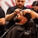 A barber is carefully trimming the hair of a customer who is leaning back in a barber's chair. The focus is on the precision and attention to detail in grooming the customer's beard and hair. The background features other barbers at work, adding to the lively atmosphere of the barbershop.