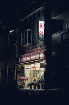 A dimly lit street scene featuring a small flower shop with a canopy and a bright illuminated sign displaying Japanese characters. Inside, various colorful plants and flowers are arranged near the entrance, creating a quaint and inviting atmosphere. The building facade is largely dark, contrasting with the warm glow emanating from the shop's interior.