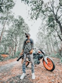 A person wearing casual clothing and a cap stands confidently beside a motorcycle with bright orange wheels, holding a camera with a large lens. The scene is set in a forest path covered with fallen leaves, surrounded by tall trees and dense foliage.