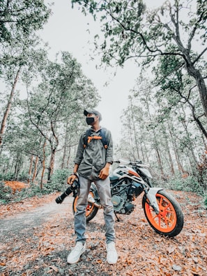 A person wearing casual clothing and a cap stands confidently beside a motorcycle with bright orange wheels, holding a camera with a large lens. The scene is set in a forest path covered with fallen leaves, surrounded by tall trees and dense foliage.