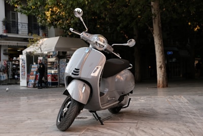 A sleek silver scooter is parked on a spacious marble pavement. In the background, there is a kiosk with various souvenirs and a tree providing shade. A person is standing near the kiosk.