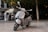 A sleek silver scooter is parked on a spacious marble pavement. In the background, there is a kiosk with various souvenirs and a tree providing shade. A person is standing near the kiosk.