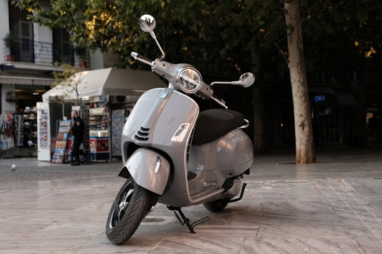 A sleek silver scooter is parked on a spacious marble pavement. In the background, there is a kiosk with various souvenirs and a tree providing shade. A person is standing near the kiosk.