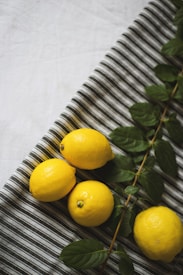 Four bright yellow lemons are placed on a striped fabric surface accompanied by a branch with glossy green leaves. The composition creates a fresh and vibrant appearance.