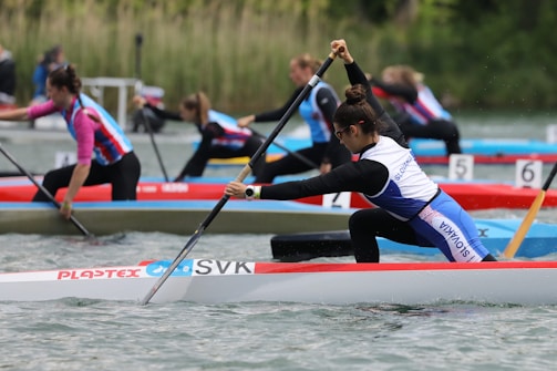Several individuals are competing in a canoeing race, paddling vigorously on a body of water. Each participant is in a separate lane, wearing athletic gear, with some competitors appearing more focused and intensely engaged in their strokes. The environment shows natural greenery in the background, suggesting an outdoor setting.