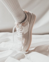 A close-up of a white sneaker with beige accents worn on a foot, paired with a ribbed high white sock, against a soft, light background.