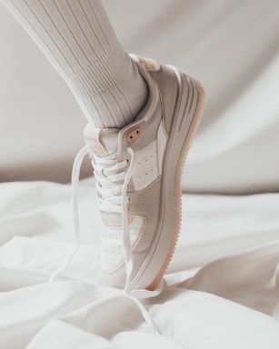 A close-up of a white sneaker with beige accents worn on a foot, paired with a ribbed high white sock, against a soft, light background.