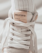 A close-up image of the upper part of a sneaker, highlighting the laces and tongue. The sneaker is made of textured gray material with white laces, and a label on the tongue reads 'PR 072819'. A white sock is visible inside the shoe.