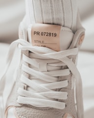 A close-up image of the upper part of a sneaker, highlighting the laces and tongue. The sneaker is made of textured gray material with white laces, and a label on the tongue reads 'PR 072819'. A white sock is visible inside the shoe.