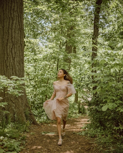 A woman in a flowing, earth-colored dress walking through a sunlit forest path.
