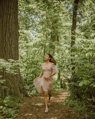 A woman in a light, flowing dress is walking along a forest path surrounded by lush green trees and foliage. Sunlight filters through the leaves, creating a dappled pattern on the path.