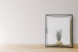 A minimalist art piece featuring a framed print of a pineapple. The artwork is encased in a geometric black frame, resting on a light wooden surface against a plain light-colored wall. The setting is simple and clean, highlighting the artistic nature of the image.