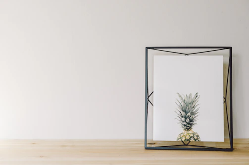 A minimalist art piece featuring a framed print of a pineapple. The artwork is encased in a geometric black frame, resting on a light wooden surface against a plain light-colored wall. The setting is simple and clean, highlighting the artistic nature of the image.