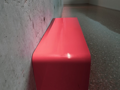 A sleek, modern bench with a smooth, curved surface is positioned against a rough-textured, gray concrete wall. The bench is colored in a vibrant shade of pink and is placed on a speckled gray floor, creating a contrast between the bright bench and the subdued surroundings. The setting appears to be an interior space, possibly a gallery or a minimalist room.