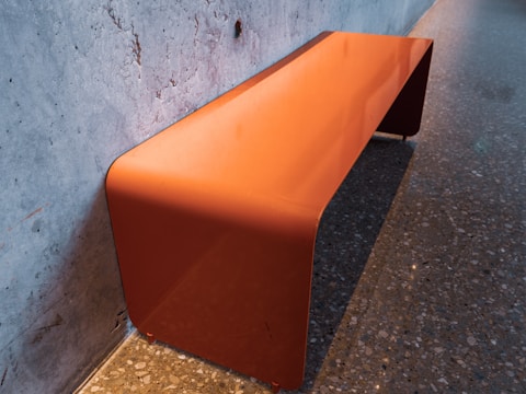 An orange metal bench is placed against a textured concrete wall. The bench has a sleek, minimalist design with smooth curves and a shiny surface. The floor is covered with speckled stone tiles, creating a contrast with the orange hue of the bench.