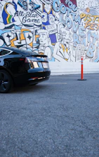 A vibrant mural featuring colorful illustrations and text related to basketball and urban themes. The mural has various elements like a jersey with the number 32, figures resembling comic characters, text like 'All-Star Game' and 'Gotta Love It'. A sleek black car is parked in front of the mural on a gray asphalt surface, with an orange traffic cone nearby.