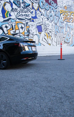 A vibrant mural featuring colorful illustrations and text related to basketball and urban themes. The mural has various elements like a jersey with the number 32, figures resembling comic characters, text like 'All-Star Game' and 'Gotta Love It'. A sleek black car is parked in front of the mural on a gray asphalt surface, with an orange traffic cone nearby.