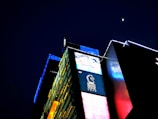 A brightly illuminated urban building with vibrant digital advertisements covers the facade. The night sky features a small, bright moon above the structure, which has panels of yellow, blue, and red lights that create a visually striking contrast against the dark background. The signage includes a logo with a horse and text below it.