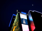 A brightly illuminated urban building with vibrant digital advertisements covers the facade. The night sky features a small, bright moon above the structure, which has panels of yellow, blue, and red lights that create a visually striking contrast against the dark background. The signage includes a logo with a horse and text below it.