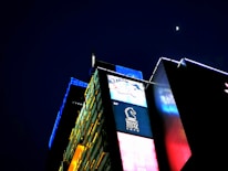 A brightly illuminated urban building with vibrant digital advertisements covers the facade. The night sky features a small, bright moon above the structure, which has panels of yellow, blue, and red lights that create a visually striking contrast against the dark background. The signage includes a logo with a horse and text below it.