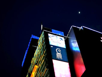 A brightly illuminated urban building with vibrant digital advertisements covers the facade. The night sky features a small, bright moon above the structure, which has panels of yellow, blue, and red lights that create a visually striking contrast against the dark background. The signage includes a logo with a horse and text below it.