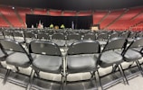 Rows of empty black chairs are neatly arranged in a large indoor arena with red stadium seating. A stage is set up at the front with a podium, potted plants, and flags. The words 'Class of' are partially visible on a sign, suggesting a graduation ceremony setting.