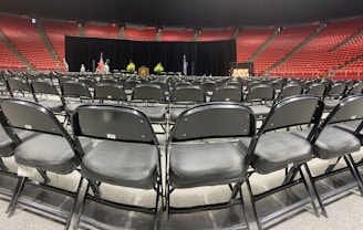 Rows of empty black chairs are neatly arranged in a large indoor arena with red stadium seating. A stage is set up at the front with a podium, potted plants, and flags. The words 'Class of' are partially visible on a sign, suggesting a graduation ceremony setting.