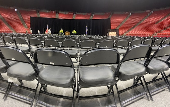 Rows of empty black chairs are neatly arranged in a large indoor arena with red stadium seating. A stage is set up at the front with a podium, potted plants, and flags. The words 'Class of' are partially visible on a sign, suggesting a graduation ceremony setting.