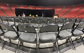 Rows of empty black chairs are neatly arranged in a large indoor arena with red stadium seating. A stage is set up at the front with a podium, potted plants, and flags. The words 'Class of' are partially visible on a sign, suggesting a graduation ceremony setting.