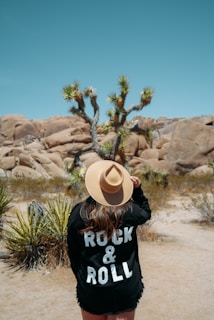 A person stands in a desert landscape wearing a wide-brimmed hat and a black jacket with 'Rock & Roll' printed on the back. Joshua trees and large rock formations are visible in the background, under a clear blue sky.