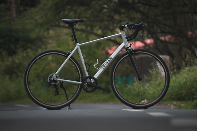 A sleek, white road bike with drop handlebars is positioned on a paved path. The brand name 'Triban' is visible on the frame. The background is filled with lush greenery, giving a serene and natural atmosphere.