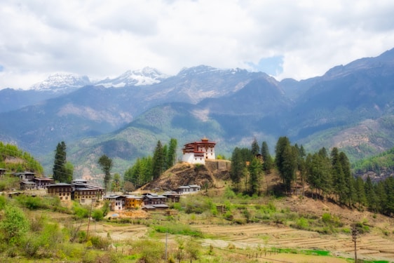 A picturesque landscape featuring a traditional Bhutanese monastery perched on a hill, surrounded by lush, green forest and rolling hills. In the background, snow-capped mountains rise majestically under a partly cloudy sky. A small village with traditional buildings spreads out at the base of the hill.
