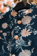 A richly decorated dark fabric garment featuring intricate floral embroidery in soft peach and pastel shades. The garment includes detailed buttons with a raised floral pattern and is suspended on a black hanger. The background contains warm, blurred foliage.