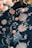A richly decorated dark fabric garment featuring intricate floral embroidery in soft peach and pastel shades. The garment includes detailed buttons with a raised floral pattern and is suspended on a black hanger. The background contains warm, blurred foliage.