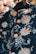 A richly decorated dark fabric garment featuring intricate floral embroidery in soft peach and pastel shades. The garment includes detailed buttons with a raised floral pattern and is suspended on a black hanger. The background contains warm, blurred foliage.