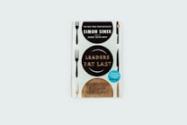 A book cover featuring the title 'Leaders Eat Last' by Simon Sinek. The design incorporates forks and knives along the edges, with informative text about the book's bestseller status and additional chapters included.