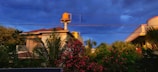 A residential area featuring a house with a tiled roof and a prominent water tank on top. Surrounding the house are lush green trees and vibrant pink flowers. Overhead, power lines stretch under a deep blue, cloudy sky.