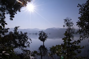 The image captures a tranquil landscape with a serene lake reflecting the sky and surrounding trees. The sun shines brightly, casting a clear, radiant light that creates a peaceful and calm atmosphere. Silhouetted trees frame the scene, and misty mountains are visible in the background.