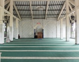 A spacious interior of a mosque with rows of green carpet, arched white marble walls, and tall pillars. The ceiling is vaulted with wooden beams, and there is a wooden podium at the front. There are people seen praying, and a digital clock on the wall shows the time. A hanging chandelier and a speaker are visible.